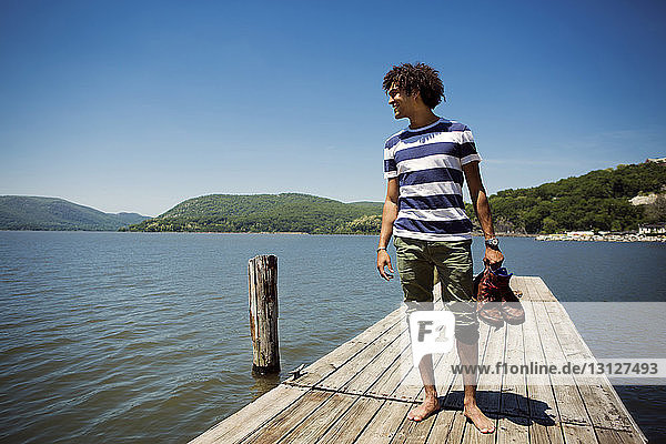 Smiling man holding shoes while standing on jetty over lake