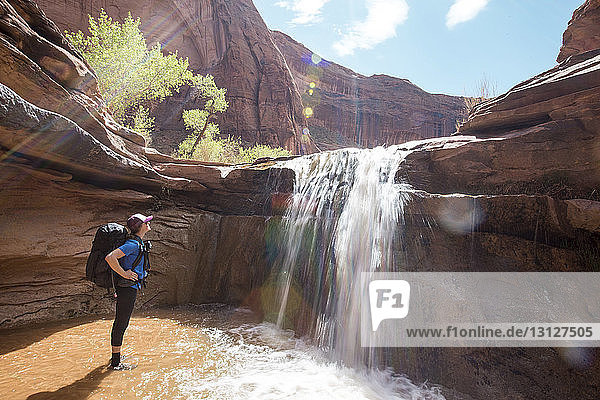 Seitenansicht einer Frau mit Rucksack beim Blick auf einen Wasserfall gegen Felsformationen bei Sonnenschein
