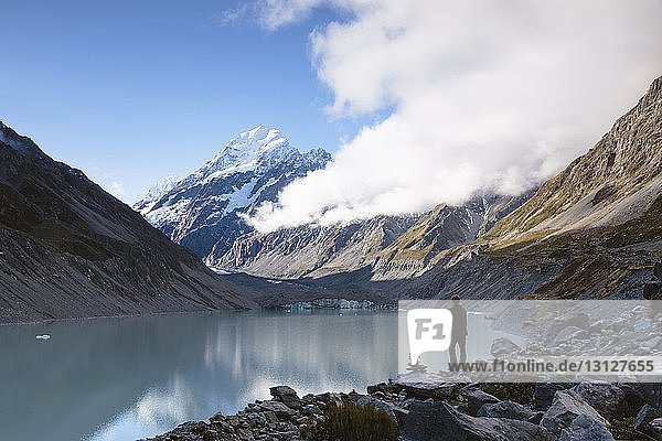 Rear view of hiker standing at lakeshore against cloudy sky at Mt Cook National Park