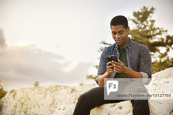 Mann benutzt Mobiltelefon  während er auf einem Felsen gegen den Himmel sitzt