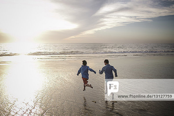 Rear view of playful brothers running at beach against sky during sunset