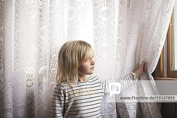 Nachdenkliches Mädchen steht vor Vorhang und schaut durchs Fenster