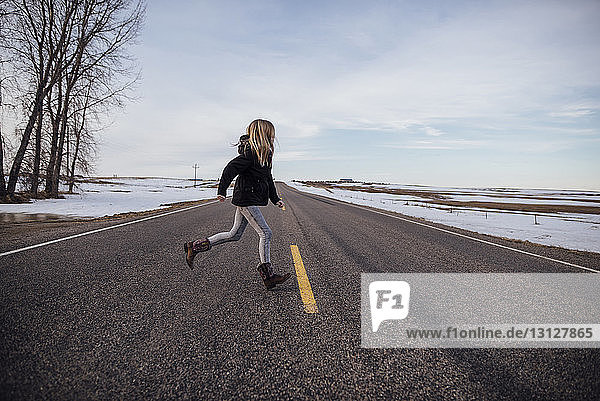Mädchen in voller Länge läuft auf der Straße inmitten eines schneebedeckten Feldes gegen den Himmel