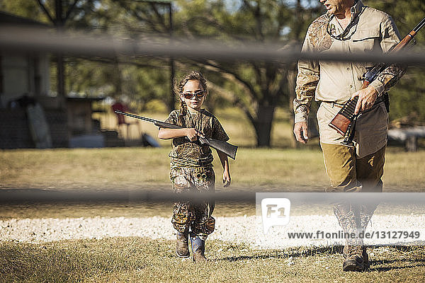 Confident girl holding rifle while walking with father on field