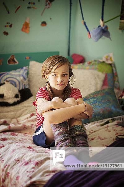 Portrait of confident girl hugging knees while sitting on bed in room