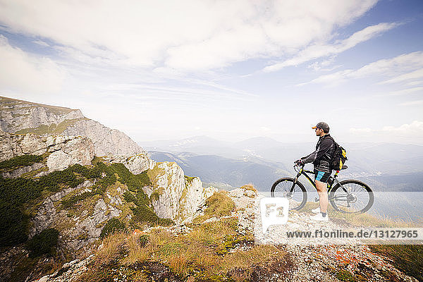 Side view of man with bicycle standing on mountain against sky