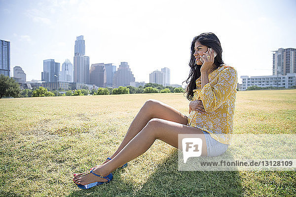 Pregnant woman using phone while sitting on grass against city