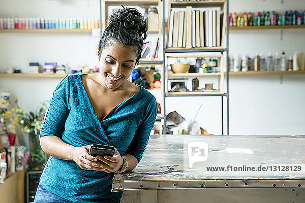 Glückliche Geschäftsfrau benutzt Smartphone  während sie sich im Büro auf den Tisch lehnt