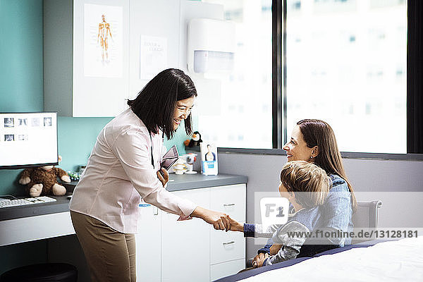 Happy female doctor greeting boy sitting with mother in clinic