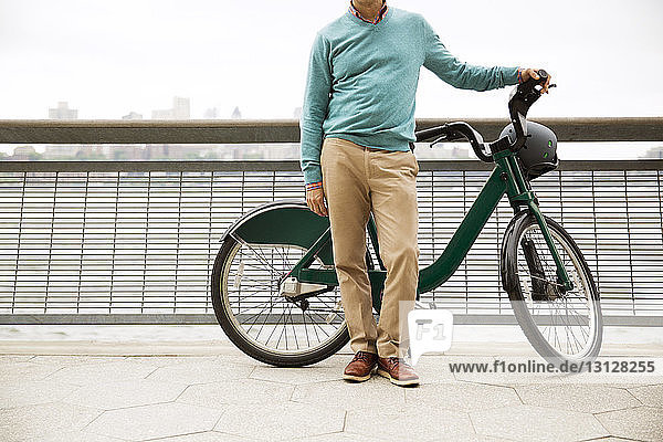 Low section of mature man standing with bicycle on promenade in city against clear sky