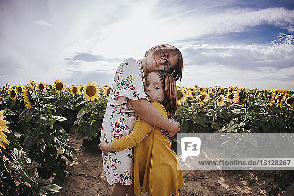 Porträt von Mädchen  die sich umarmen  während sie im Sonnenblumenfeld vor dem Himmel stehen