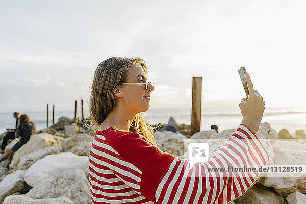 Seitenansicht einer jungen Frau  die sich am Strand mit einem Smartphone selbstständig macht