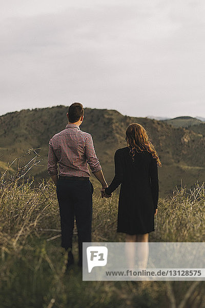 Rear view of young couple holding hands while standing on grassy field against mountains and sky