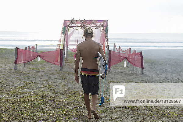 Rückansicht eines Mannes ohne Hemd mit Surfbrett  der am Strand vor klarem Himmel spazieren geht