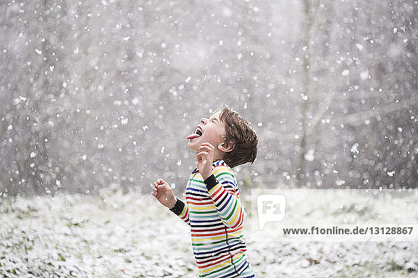 Seitenansicht eines glücklichen Jungen  der bei Schneefall die Zunge herausstreckt  während er auf dem Feld steht