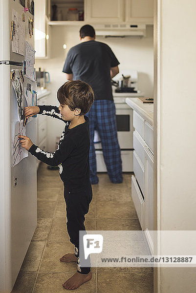Boy sticking paper on refrigerator while father working in background at kitchen