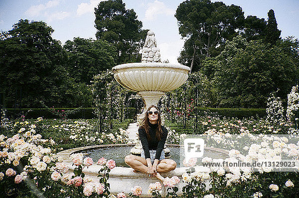 Porträt einer Frau  die auf einem von Rosen umgebenen Brunnen im Park sitzt