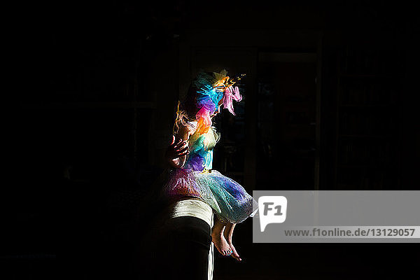 Side view of girl wearing colorful unicorn costume sitting on sofa in darkroom at home