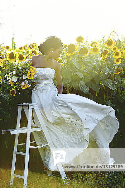 Braut hält Brautstrauss  während sie an der Leiter inmitten von Sonnenblumen am Bauernhof gegen den Himmel steht