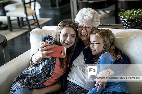 Schrägaufnahme einer glücklichen Großmutter und Enkelinnen  die auf dem Sofa sitzend per Smartphone Selbstgespräche führen
