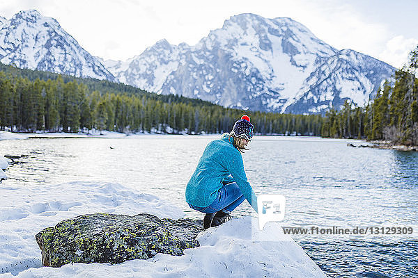 Frau in voller Länge kauert auf einem Felsen inmitten eines zugefrorenen Sees vor Bergen
