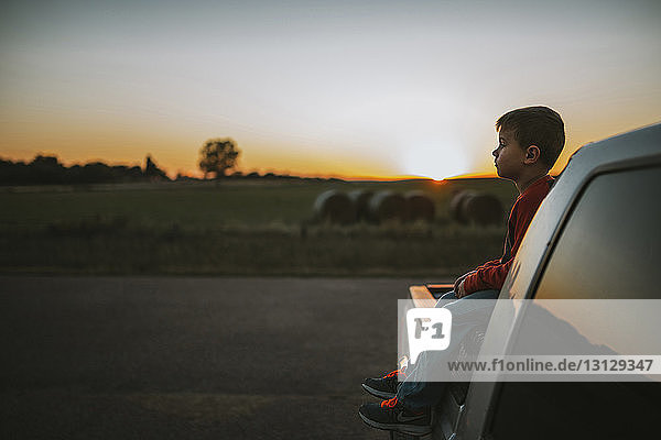 Side view of boy looking away while sitting on pick-up truck during sunset