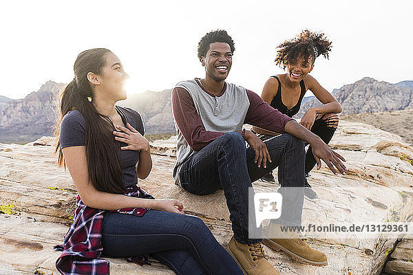 Happy friends talking while sitting on rock formations against clear sky