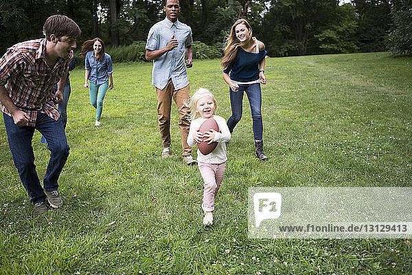 Spielerische Freunde rennen hinter einem Mädchen her  das auf einem Rasenplatz einen Fussball hält