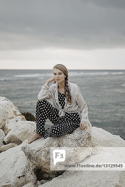 Ganzfigur einer Frau  die auf Felsen am Strand vor bewölktem Himmel sitzt