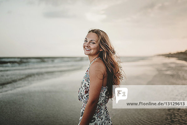 Portrait of happy woman standing at beach against sky during sunset