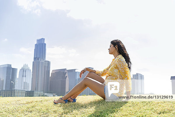 Seitenansicht einer nachdenklichen Frau  die auf Gras an Gebäuden sitzt