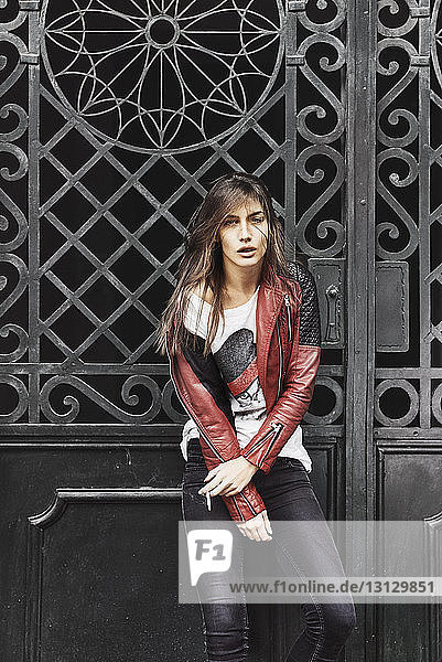 Porträt einer selbstbewussten jungen Frau in Lederjacke  die eine Zigarette gegen ein geschlossenes Metalltor hält