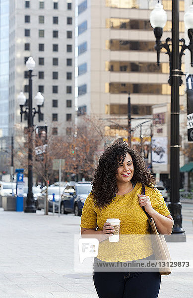 Lächelnde Frau hält Einwegbecher  während sie am Bürgersteig in der Stadt steht