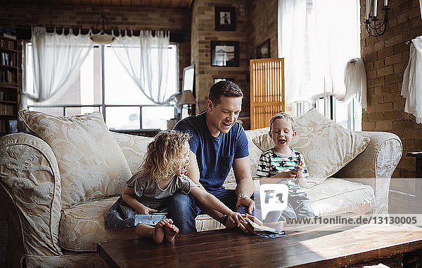 Glücklicher Vater spielt mit Kindern  während er auf dem Sofa im Wohnzimmer sitzt