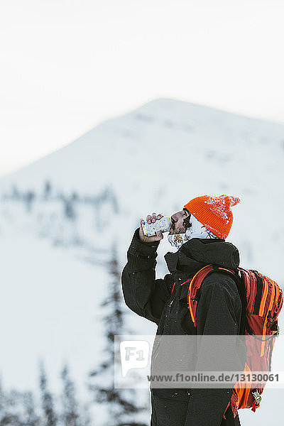 Seitenansicht eines Wanderers mit Rucksack  der aus einer Dose trinkt  während er vor einem schneebedeckten Berg steht