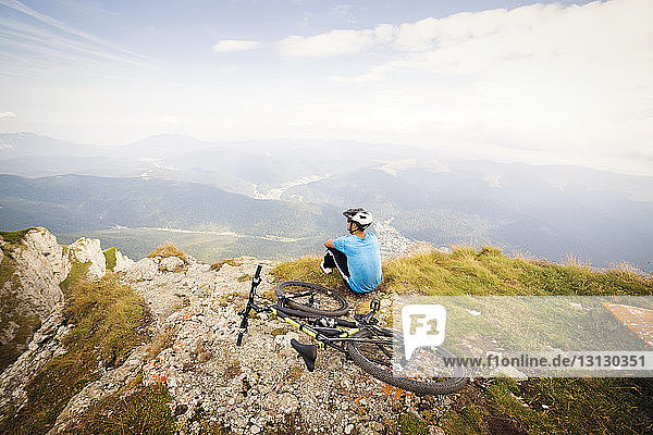 Rear view of man sitting by bicycle on mountain against sky