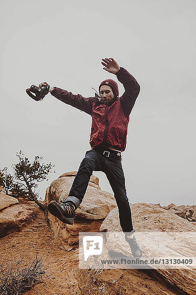 Low angle view of man holding camera while jumping on rocks against sky