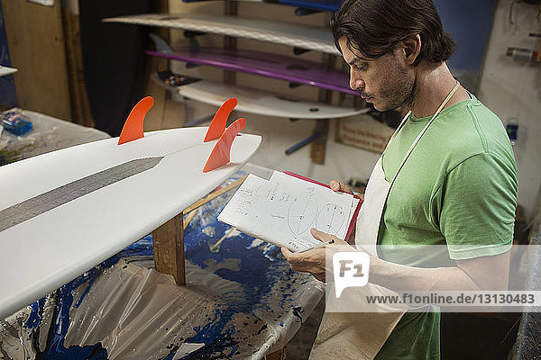 Männlicher Arbeiter betrachtet Surfbrett-Diagramme in einem Buch  während er in der Werkstatt steht