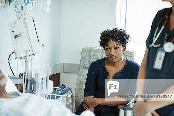 Nachdenkliche Tochter sitzt bei der Krankenschwester  während sie den Vater auf der Krankenhausstation liegen sieht
