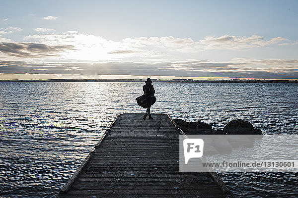 Rückansicht einer in eine Decke gehüllten Frau  die bei Sonnenuntergang auf einem Pier am Simcoe-See gegen den Himmel steht
