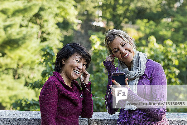Smiling female friends listening music while standing against retaining wall at park