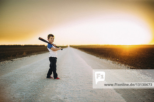 Portrait of boy holding baseball bat while standing on road amidst field against sky during sunset