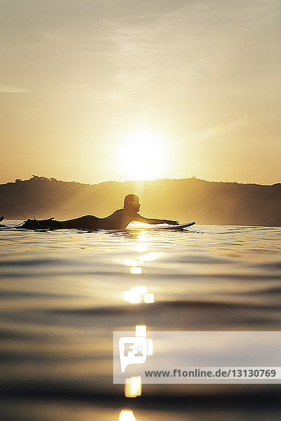 Seitenansicht einer Surferin  die bei Sonnenuntergang auf einem Surfbrett im Meer liegt