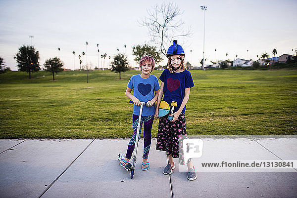 Porträt von Schwestern mit Skateboard und Schubsroller  die auf einem Fußweg gegen den Himmel im Park stehen