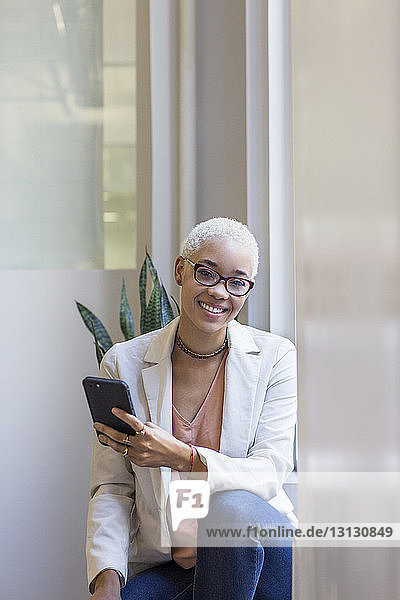Portrait of smiling woman using mobile phone while standing by window in office