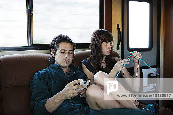 Mann benutzt Mobiltelefon  während die Frau im Wohnmobil auf dem Sofa strickt