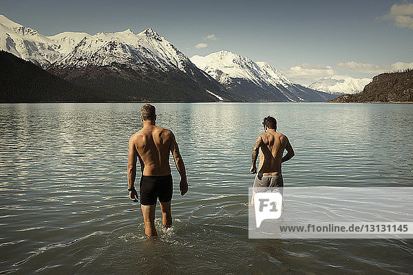 Rear view shirtless male friends walking into Kenai lake against snowcapped mountains