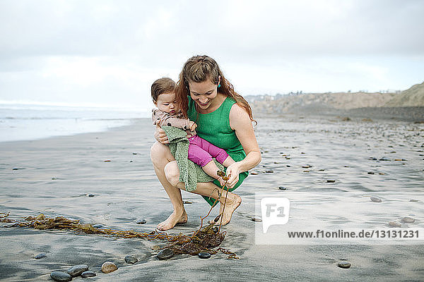 Woman with daughter touching seaweed at beach against cloudy sky