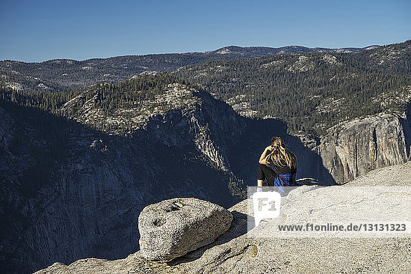 Side view of woman sitting on cliff at Yosemite National Park against blue sky