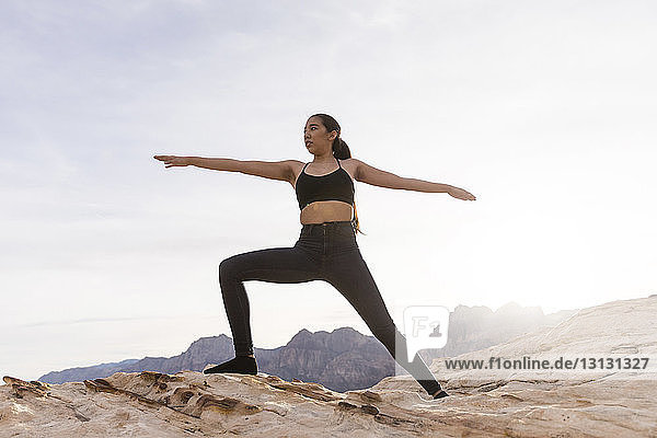 Woman practicing warrior pose on rock formation against sky